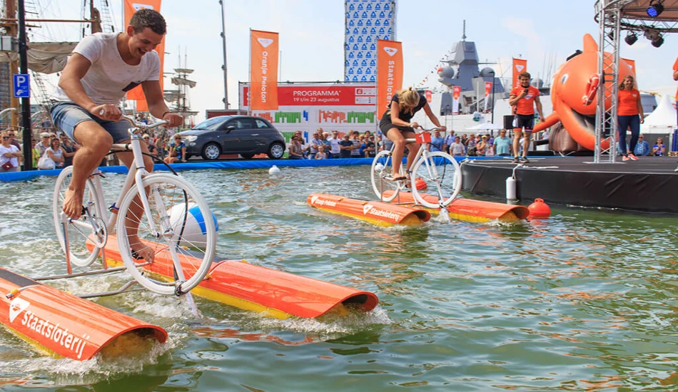 Two cyclists ride on floating platforms in a pool, surrounded by an audience and festival booths.