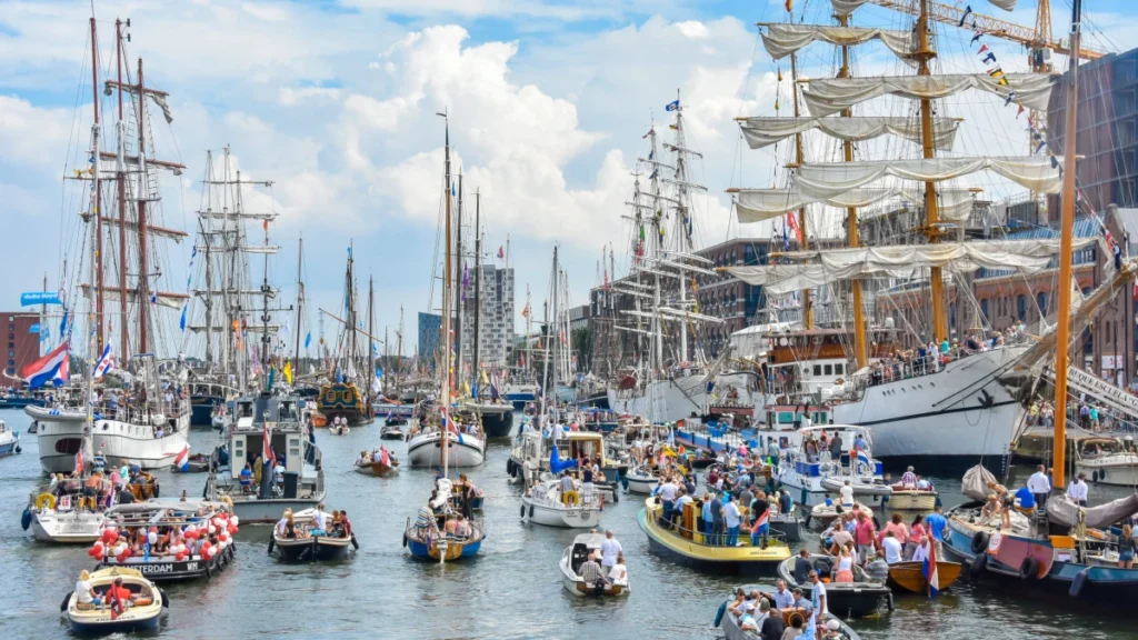 Tall-ships-surrounded-by-small boats Bustling harbor with vintage sailing ships and boats during Sail Amsterdam festival.