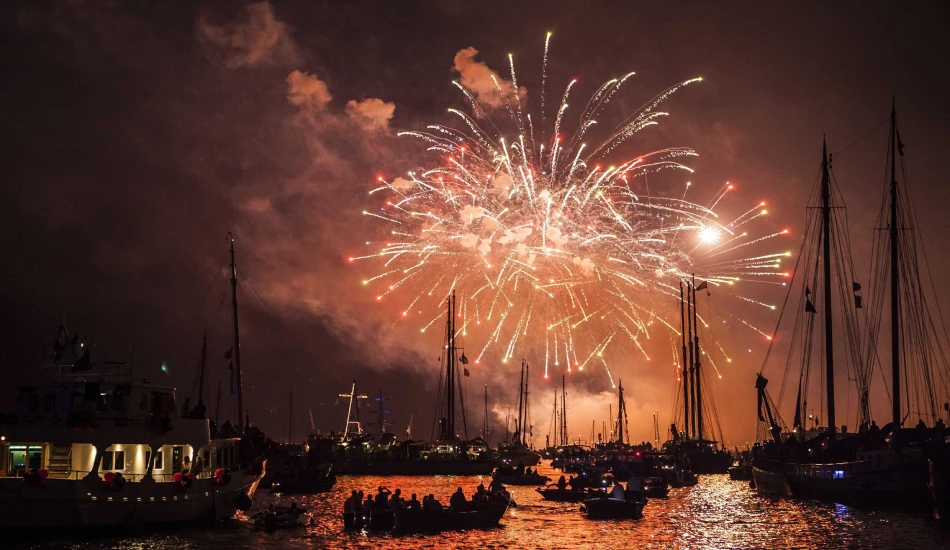 Fireworks light up the sky above tall ships and boats during Sail Amsterdam at night.