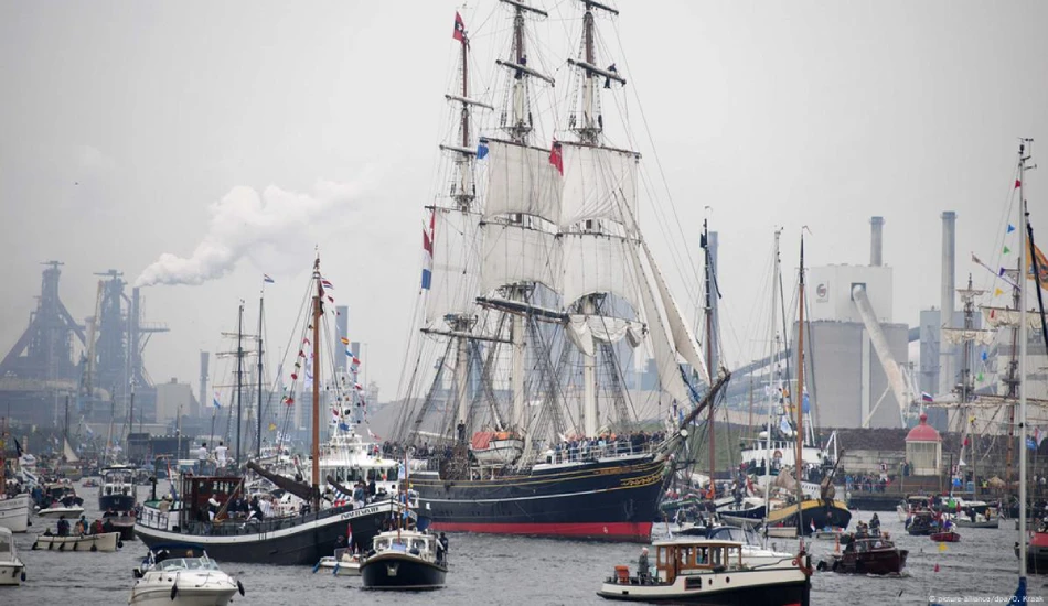 Tall ships are surrounded by small boats and cruise in a river. 