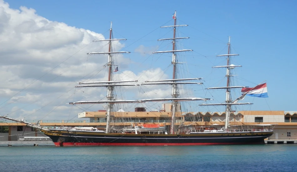 A tall ship with black and red hull docked at a harbor, flying the Dutch flag.